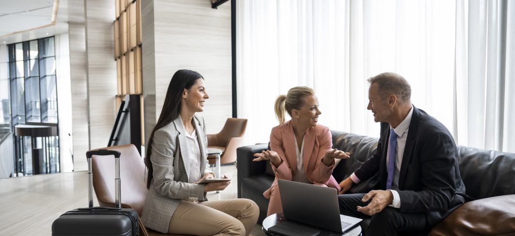 Three business colleagues seen in a hotel lobby sitting and talking while preparing for a next meeting and looking at a smart device and a lap top during their business trip.